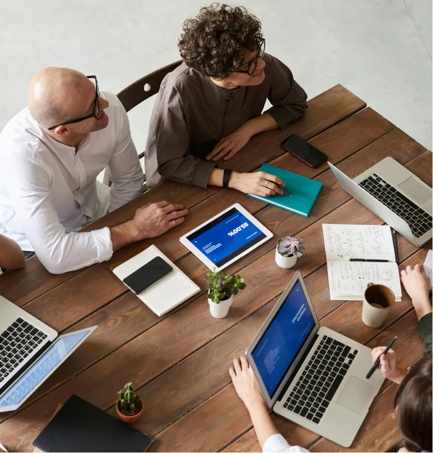 Four people sit around a wooden table with laptops, notebooks, coffee, and plants, having a meeting.